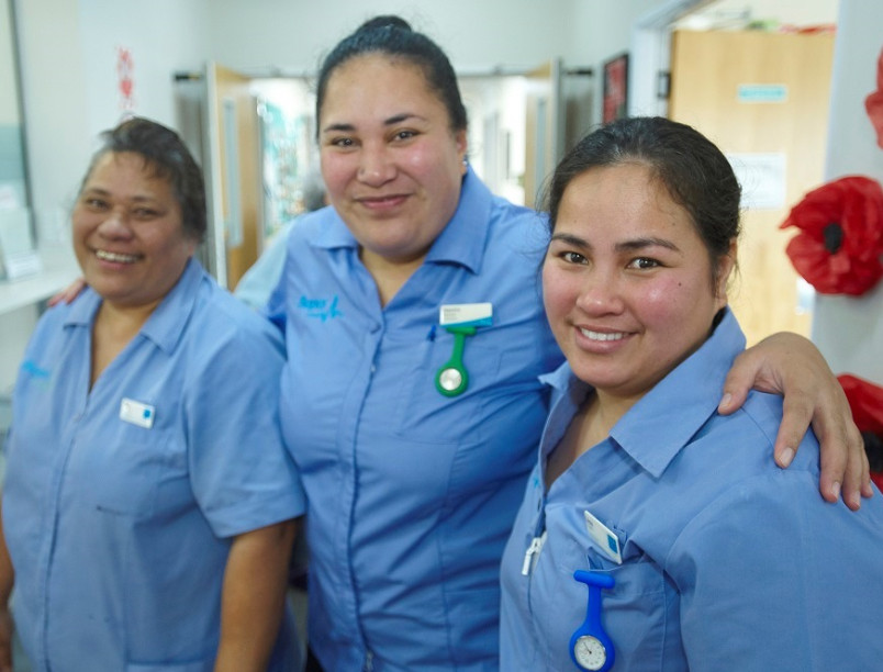 3 migrant aged care nurses standing in a rest home corridor - arms around each other and smiling.