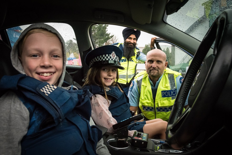 Children wearing police uniform hat and vest sitting in a car with two police men in background smiling