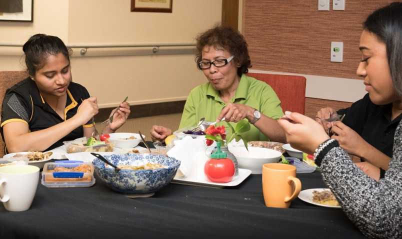 Three migrant care workers sharing homemade lunch at work in retirement village meal room