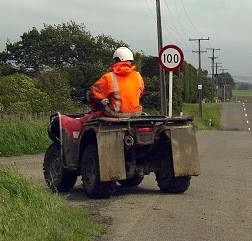 Dairy farm worker on a farm bike driving off a road into a driveway, just before a 100 k speed sign